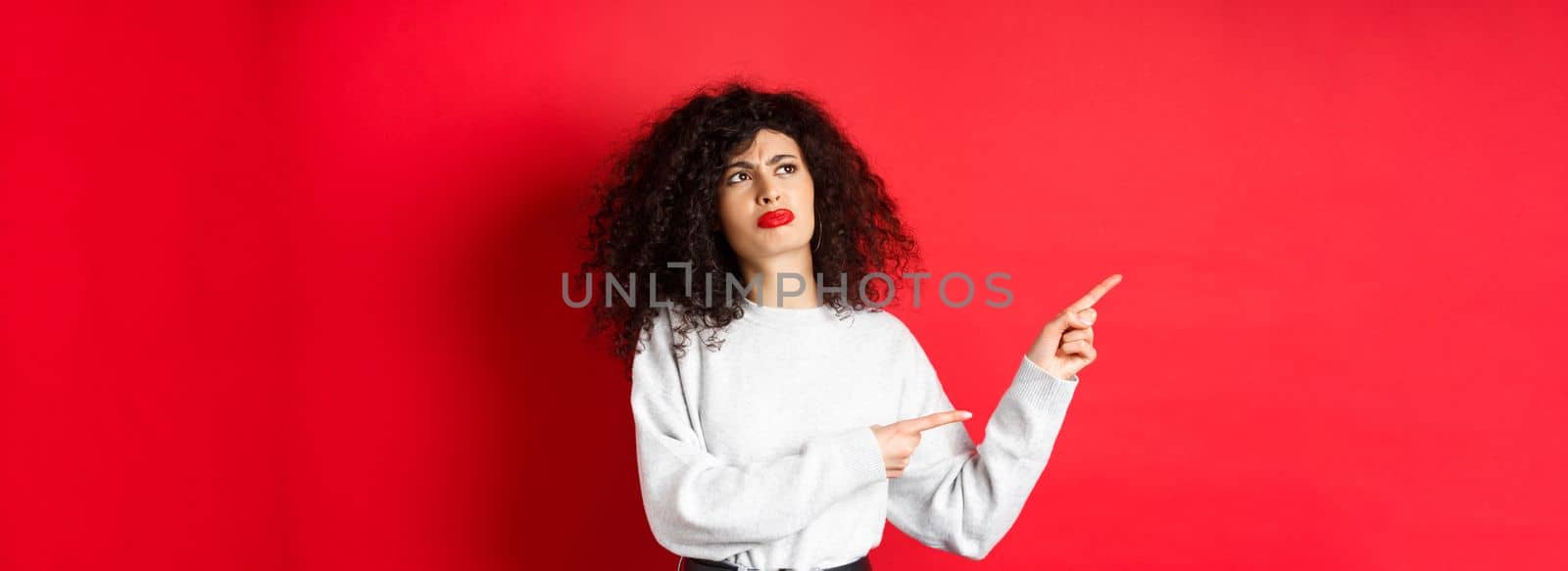 Skeptical frowning girl with curly hair, pointing and looking left hesitant, standing upset on red background.