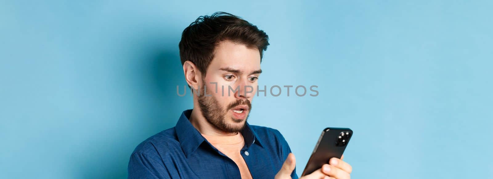 Close-up of man looking shocked and reading message on smartphone, gasping and stare startled at screen, standing on blue background.