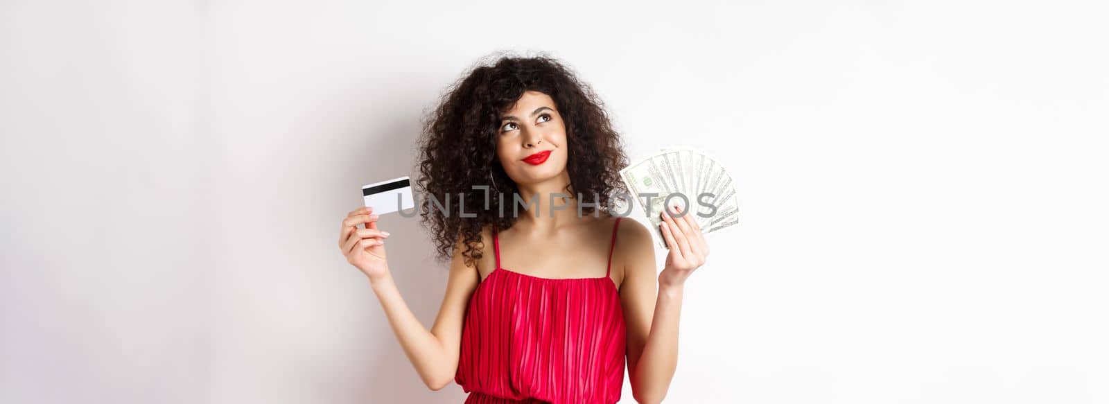 Shopping. Woman thinking and smiling, holding money with plastic credit card, wearing red elegant dress and evening makeup, white background.