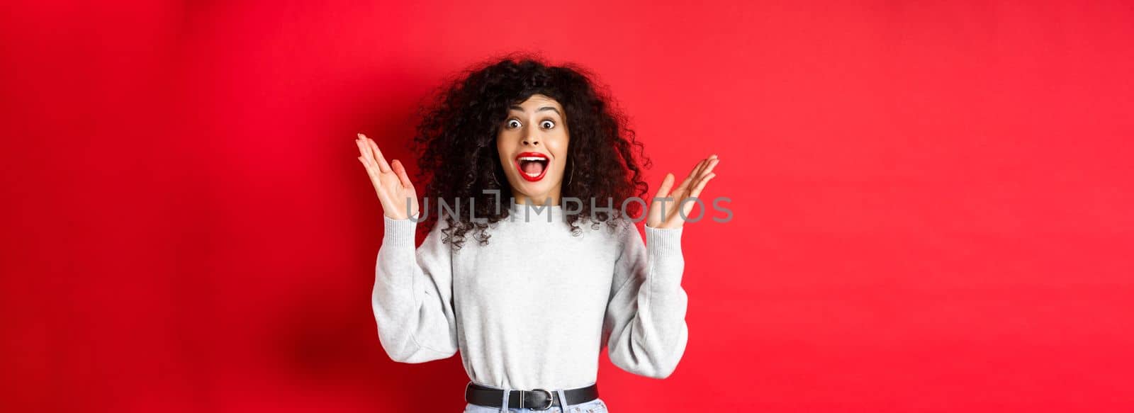 Image of happy and surprised curly woman in makeup and sweatshirt, raising hands up and rejoicing from good news, standing on red background by Benzoix