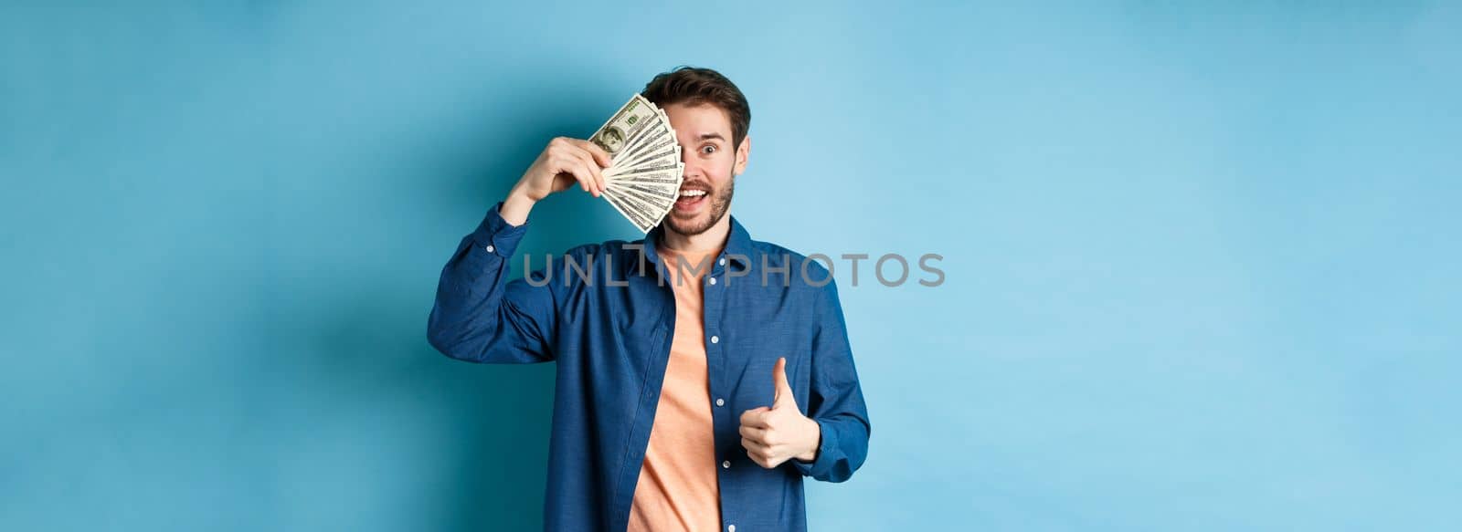 Cheerful guy cover half of face with money and showing thumbs-up, recommending fast cash loan, standing on blue background by Benzoix