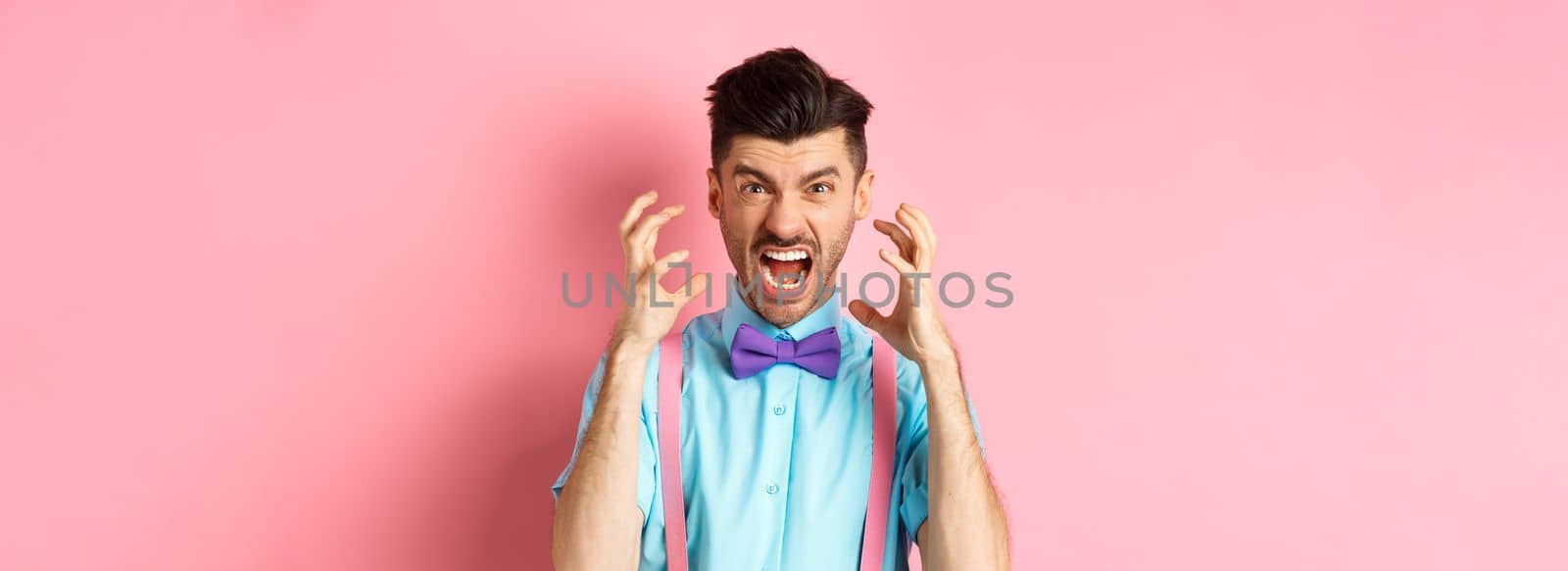 Frustrated young man losing temper, screaming and looking angry at camera, shaking hands pissed-off, standing mad on pink background.