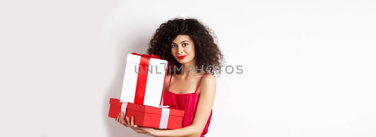 Elegant woman holding Valentines day gifts and smiling romantic at camera, standing in red dress on white background.