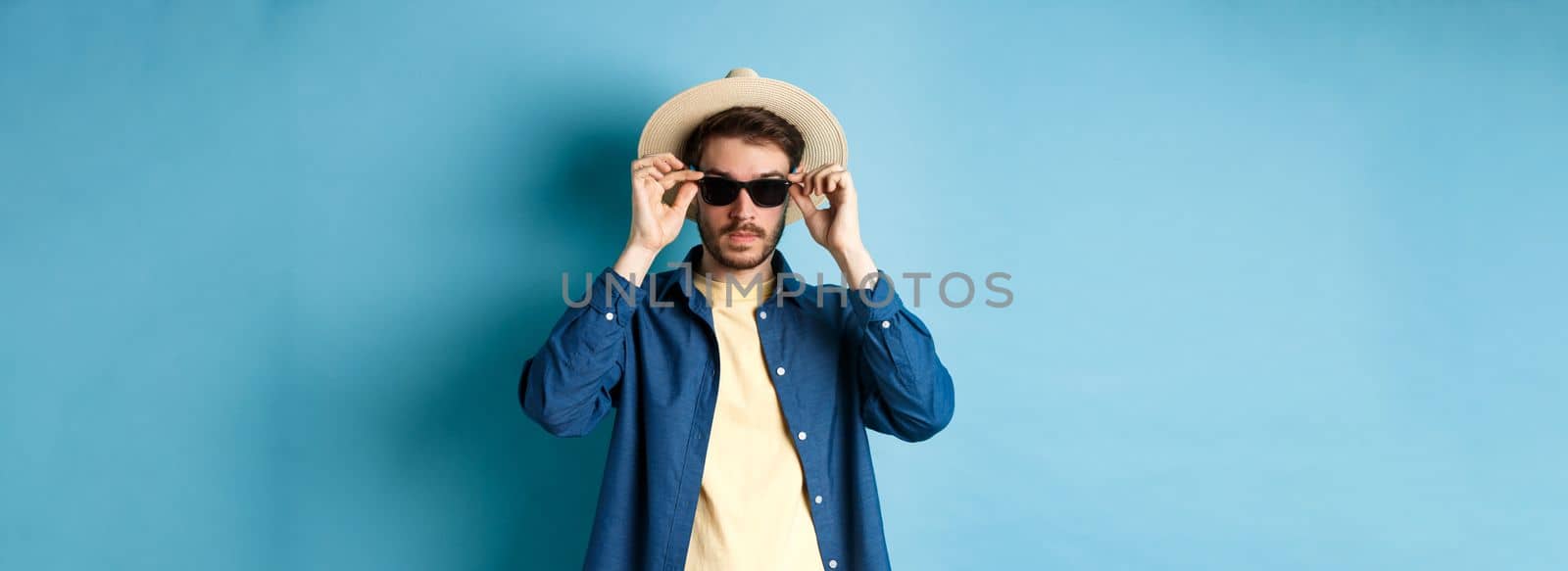 Handsome tourist in straw hat put on sunglasses on summer vacation, standing on blue background.