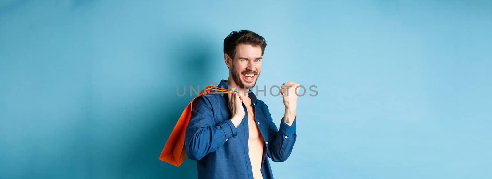 Cheerful guy saying yes, raising fist pump and smiling, holding orange shopping bag, feeling joy after buying with discounts, standing on blue background.