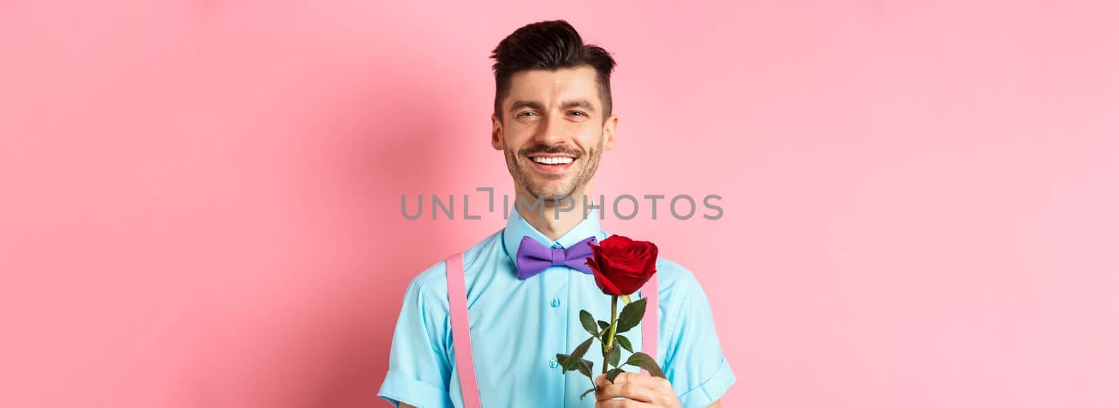 Attractive fancy guy waiting for date on lovers date, holding red rose and smiling, standing on romantic pink background. Valentines day concept by Benzoix