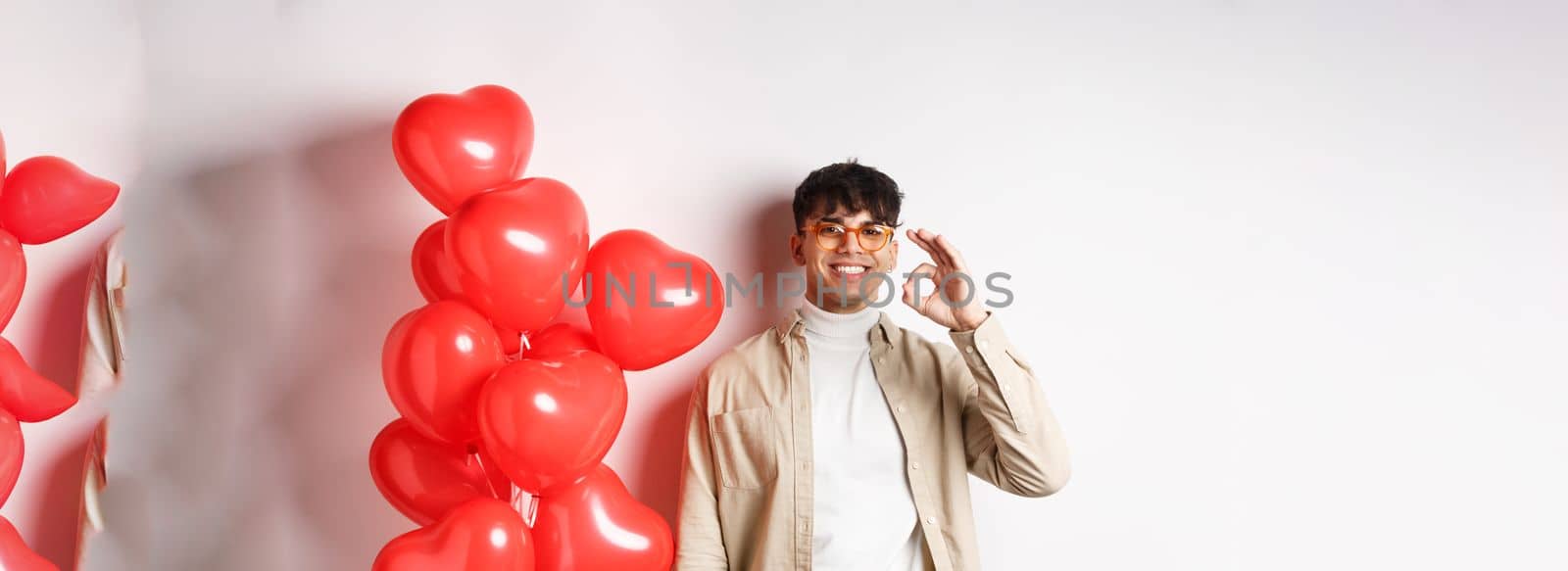 Valentines and romance concept. Happy young man smiling, showing OK sign near red hearts gesture, recommending good offer on lovers day by Benzoix
