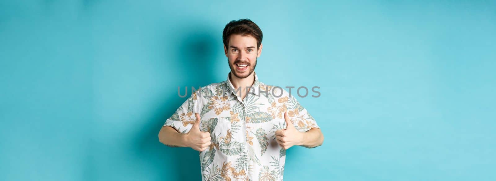 Handsome tourist showing thumbs up and say yes, praising good tourism agency, wearing hawaiian shirt, standing on blue background.