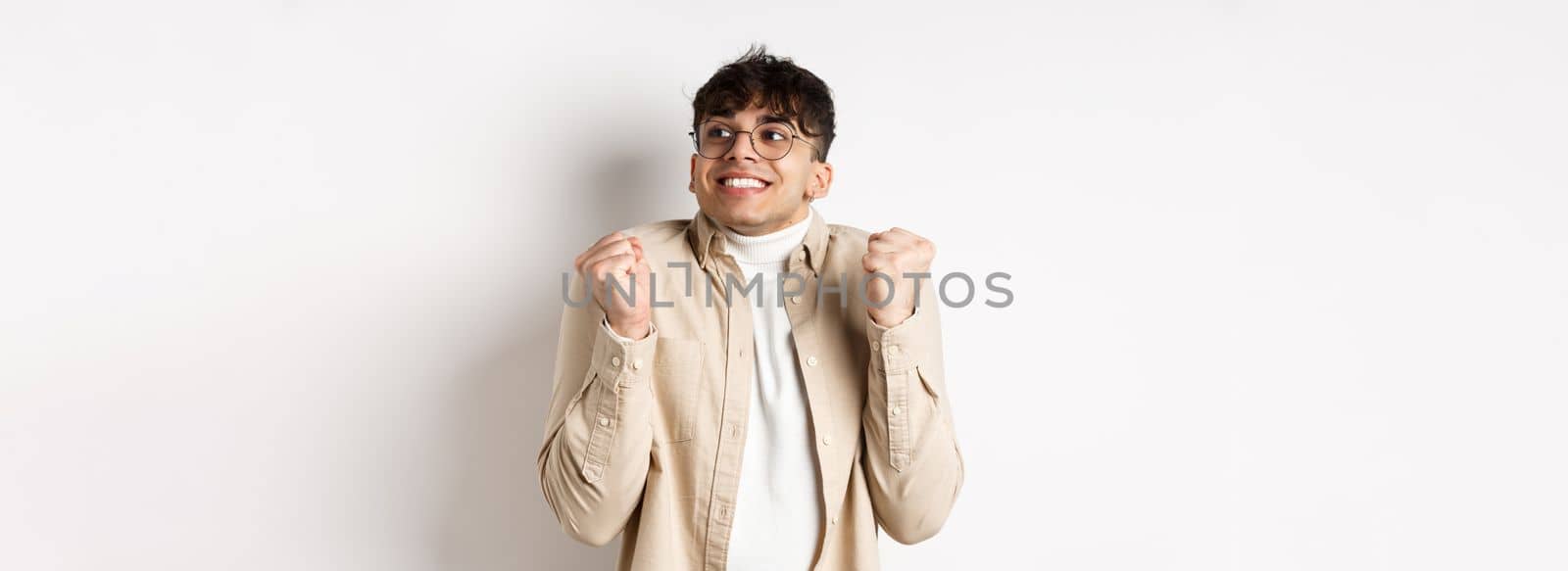 Image of handsome excited man feeling motivated and lucky, looking right and smiling, making fist pump gesture to celebrate victory, winning prize, standing on white background.