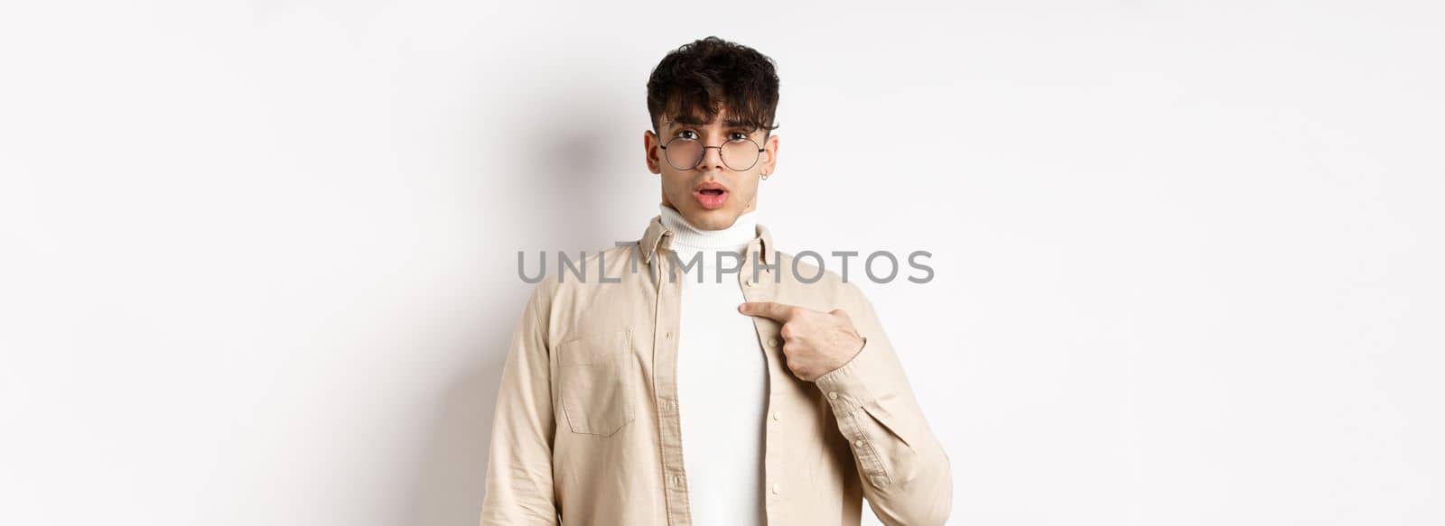 Confused and surprised young man pointing at himself, being chosen or accused, standing on white background.