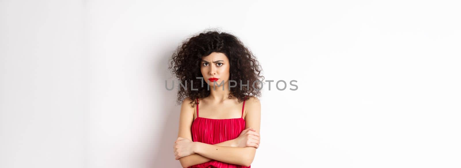 Sad and offended caucasian woman in red dress frowning, cross arms on chest and sulking, feeling mad at you, standing over white background.