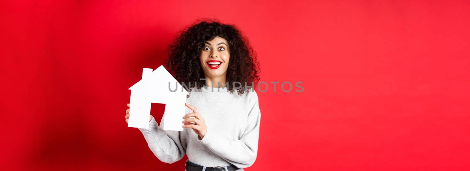 Real estate. Excited smiling woman showing paper house cutout and looking amazed, standing on red background.