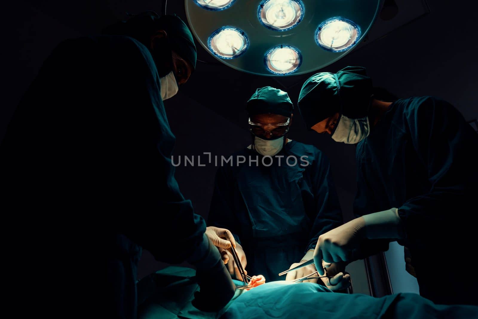 Surgical team performing surgery to patient in sterile operating room. In a surgery room lit by a lamp, a professional and confident surgical team provides medical care to an unconscious patient.