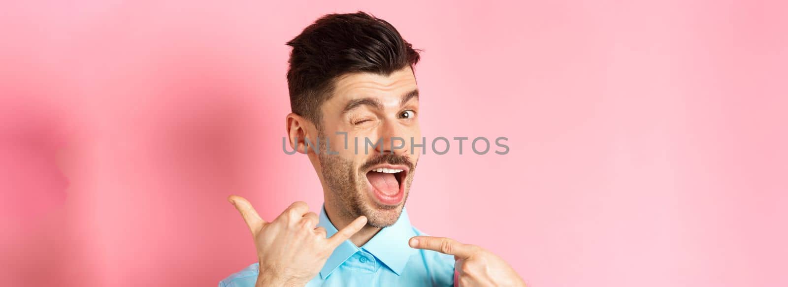 Cheeky and funny guy showing phone gesture, asking to call him and winking, telling dial his number, standing over pink background.