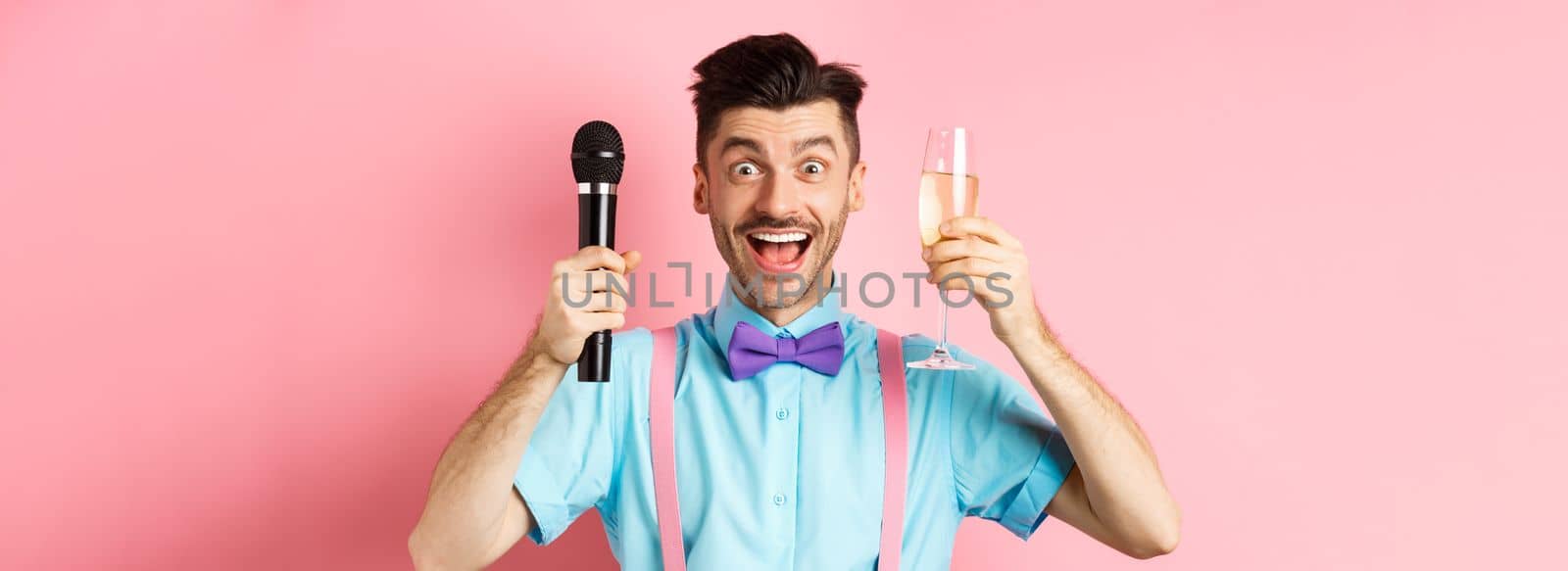 Party and festive events concept. Cheerful young male entertainer, giving speech on holiday, raising glass of chamapgne and holding microphone, making toast on wedding, pink background.
