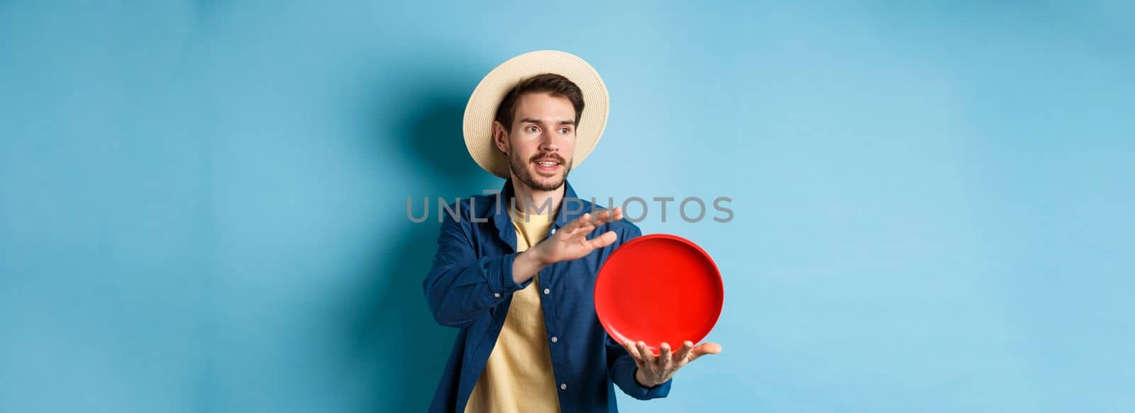 Happy smiling guy throwing frisbee, looking aside at friend, standing on blue background in straw hat by Benzoix