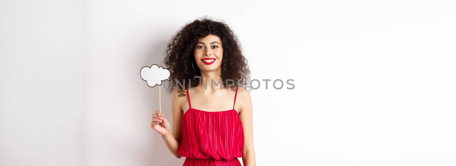 Happy stylish woman with curly hair, beauty makeup, holding comment cloud on stick and smiling, standing in red dress on white background.