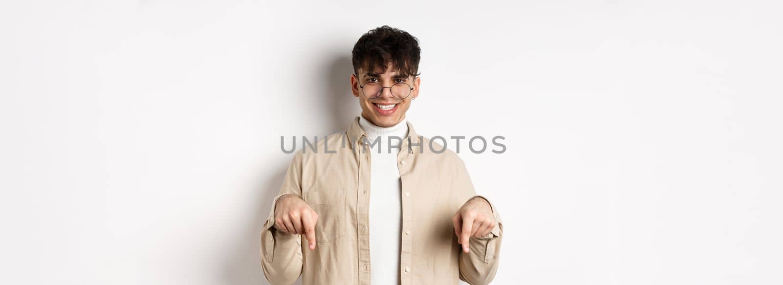 Stylish hipster male model in glasses pointing fingers down, smiling pleased at camera, recommending product, standing on white background.