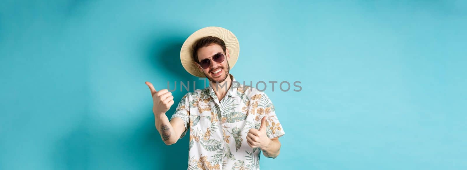 Cheerful tourist having fun summer holidays, showing thumbs up and smiling, standing in hawaiian shirt and sunglasses on blue background by Benzoix