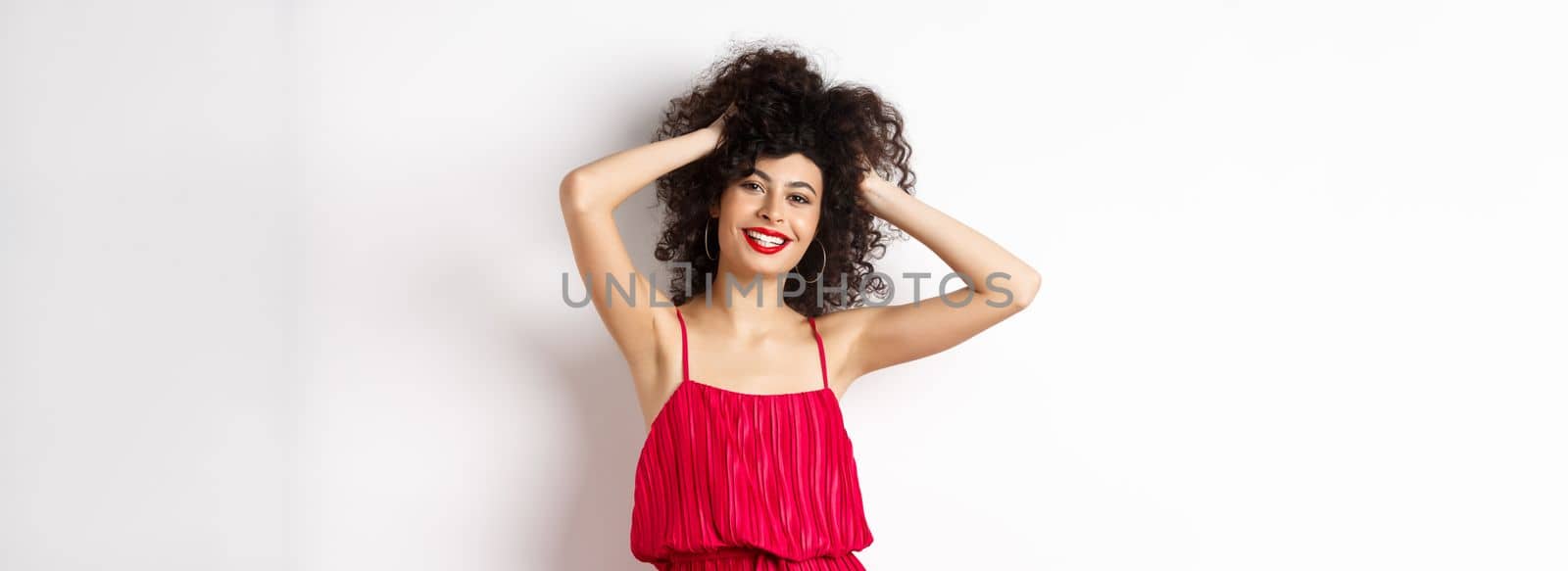 Happy smiling woman touching her curly hair and laughing, standing in red dress on white background.