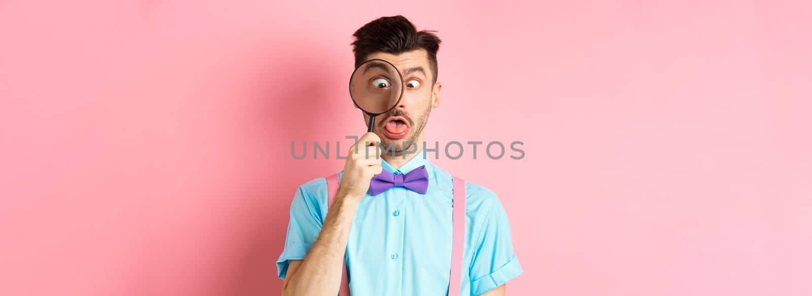 Funny man in bow-tie look through magnifying glass, squinting and making silly faces, standing on pink background by Benzoix