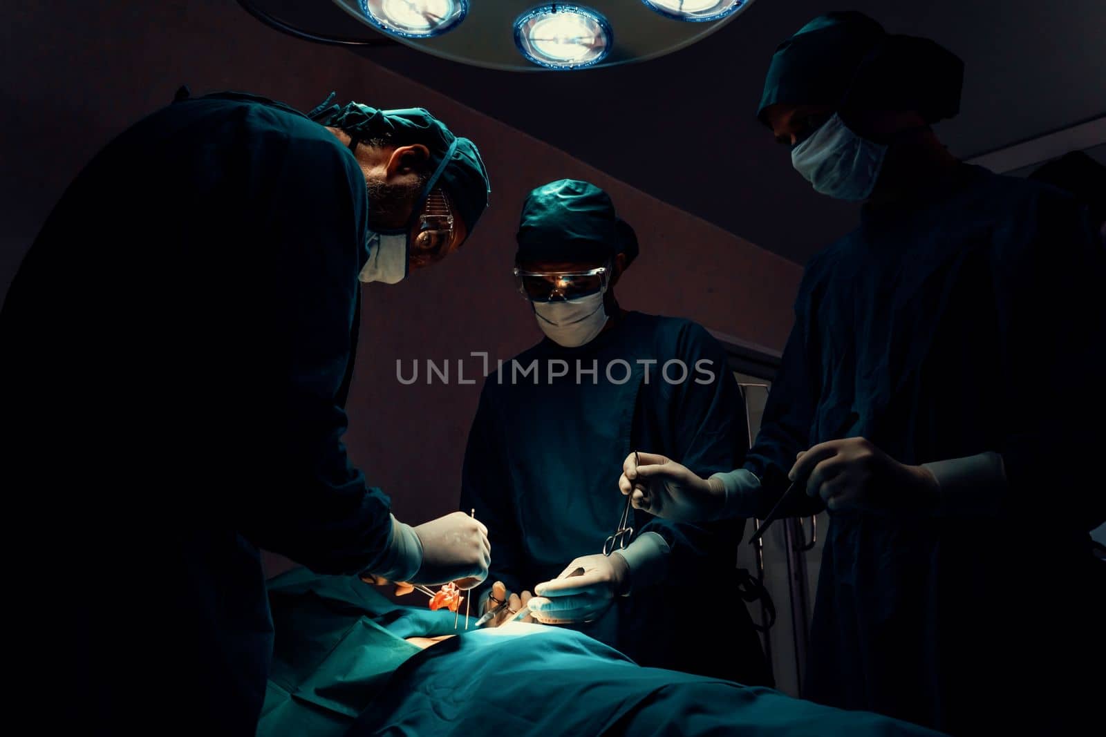 Surgical team performing surgery to patient in sterile operating room. In a surgery room lit by a lamp, a professional and confident surgical team provides medical care to an unconscious patient.