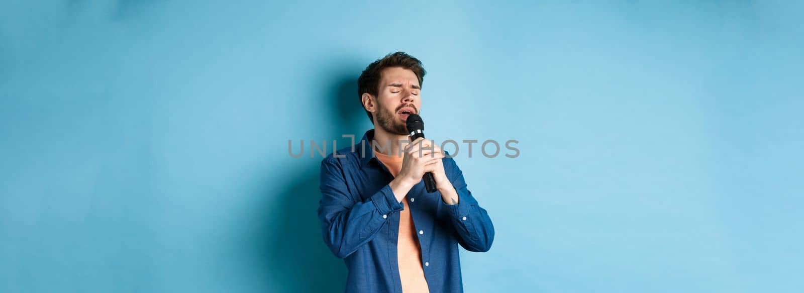 Romantic guy singing in microphone, performing on stage, standing on blue background.