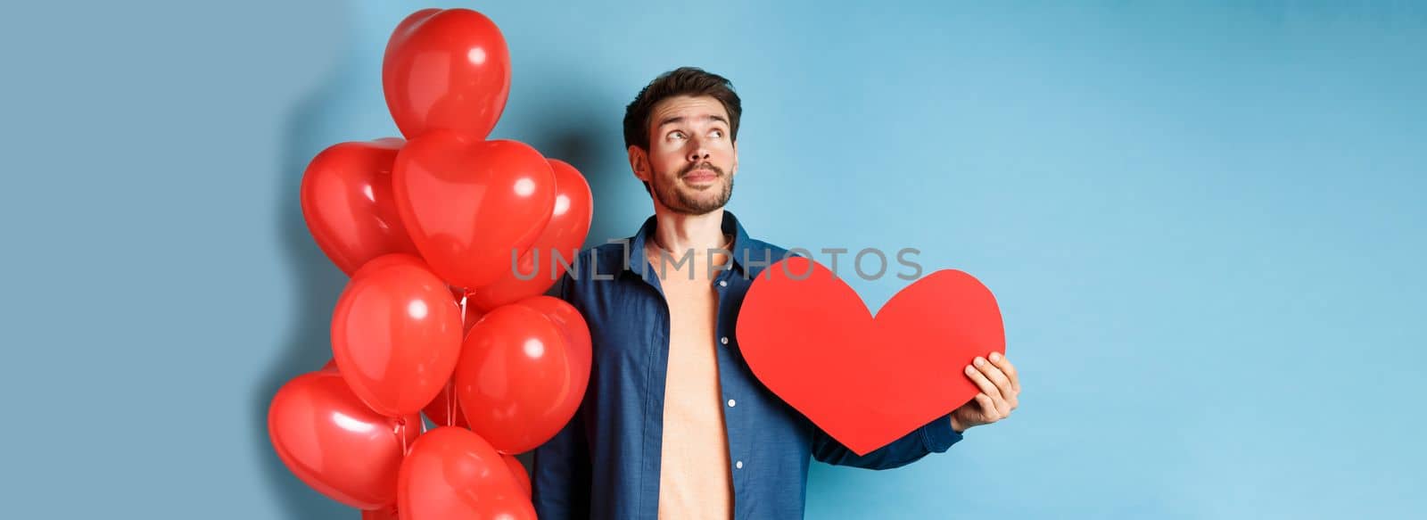 Valentines day and love concept. Man dreaming of soulmate, holding big red heart cutout and balloons, standing over blue background.