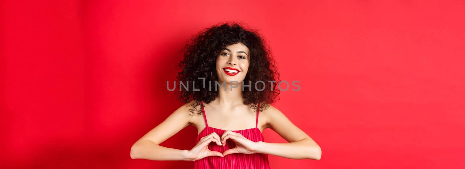 Lovers day. Beautiful woman celebrating valentines, showing heart sign and smiling, standing in romantic red dress on studio background.