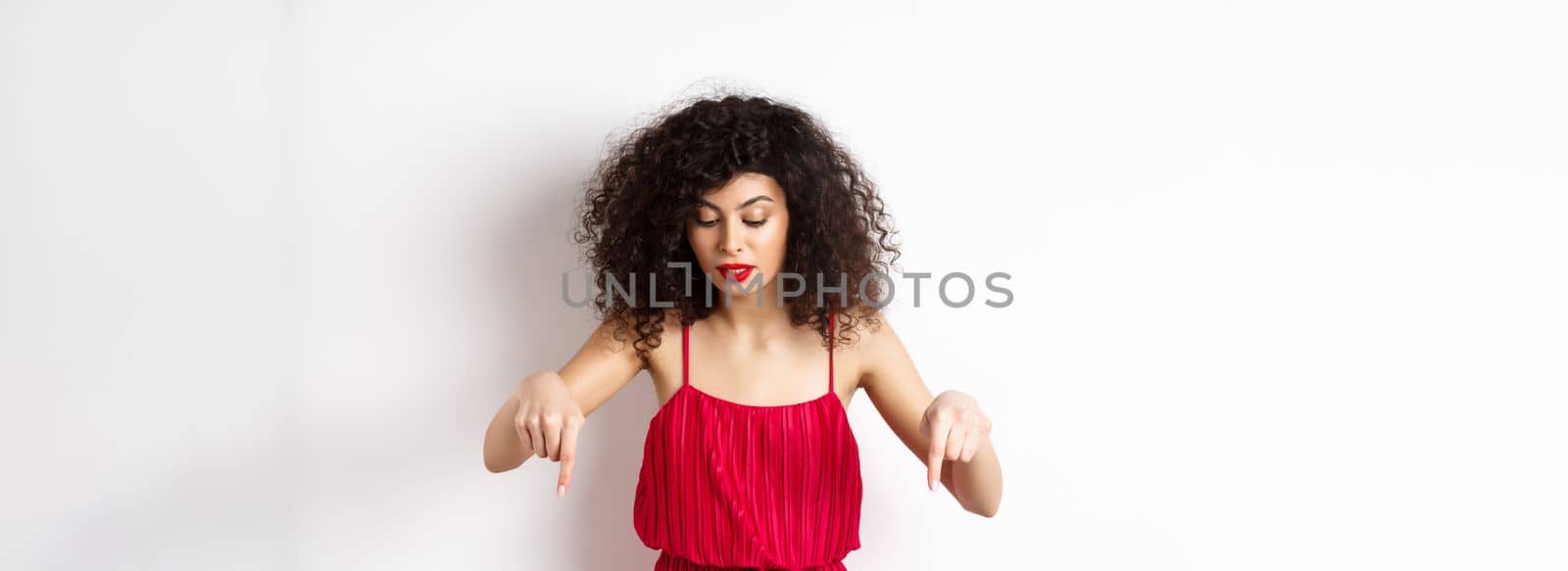 Intrigued beautiful woman with red lips and dress, pointing and looking down at banner, reading logo, standing over white background by Benzoix