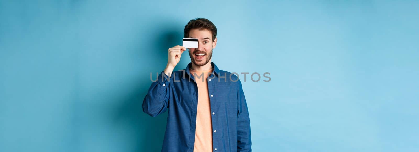 Cheerful handsome man showing plastic credit card and looking excited, checking out special offer, standing on blue background.