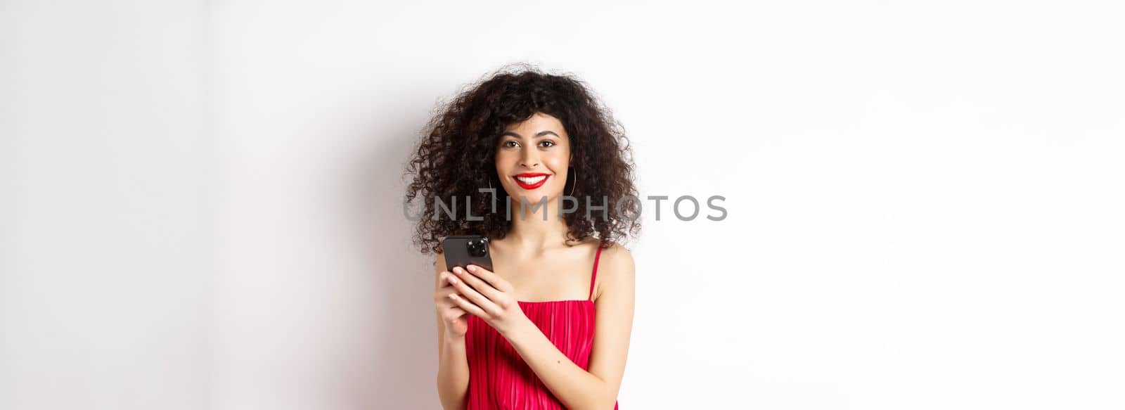 Cheerful woman with curly hair, using smartphone in red dress, smiling at camera, standing over white background. Copy space