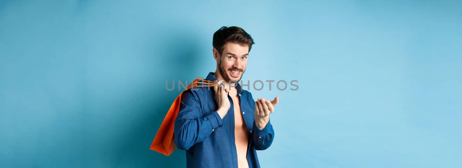 Modern guy holding shopping bag behind shoulder and beckon you, telling to come closer or inviting to store, standing on blue background.