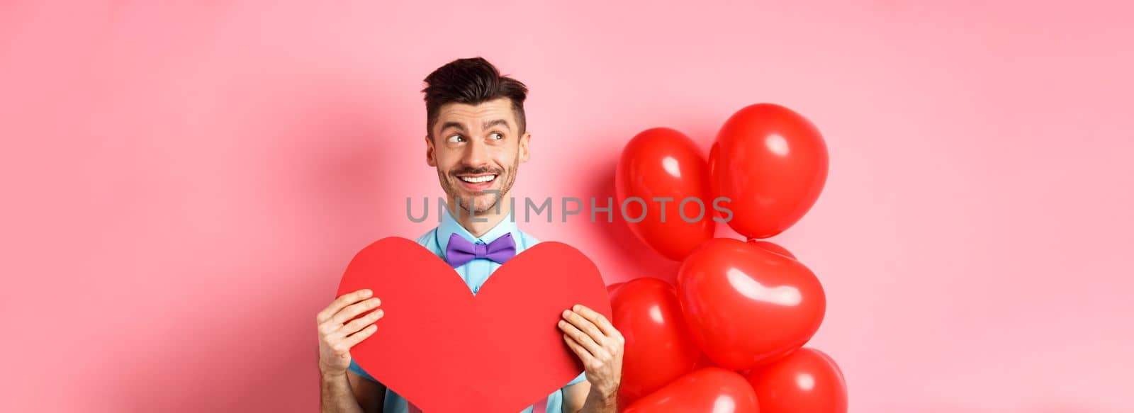 Valentines day concept. Romantic guy smiling and looking left, dreaming of date with lover, showing red big heart cutout, pink background.