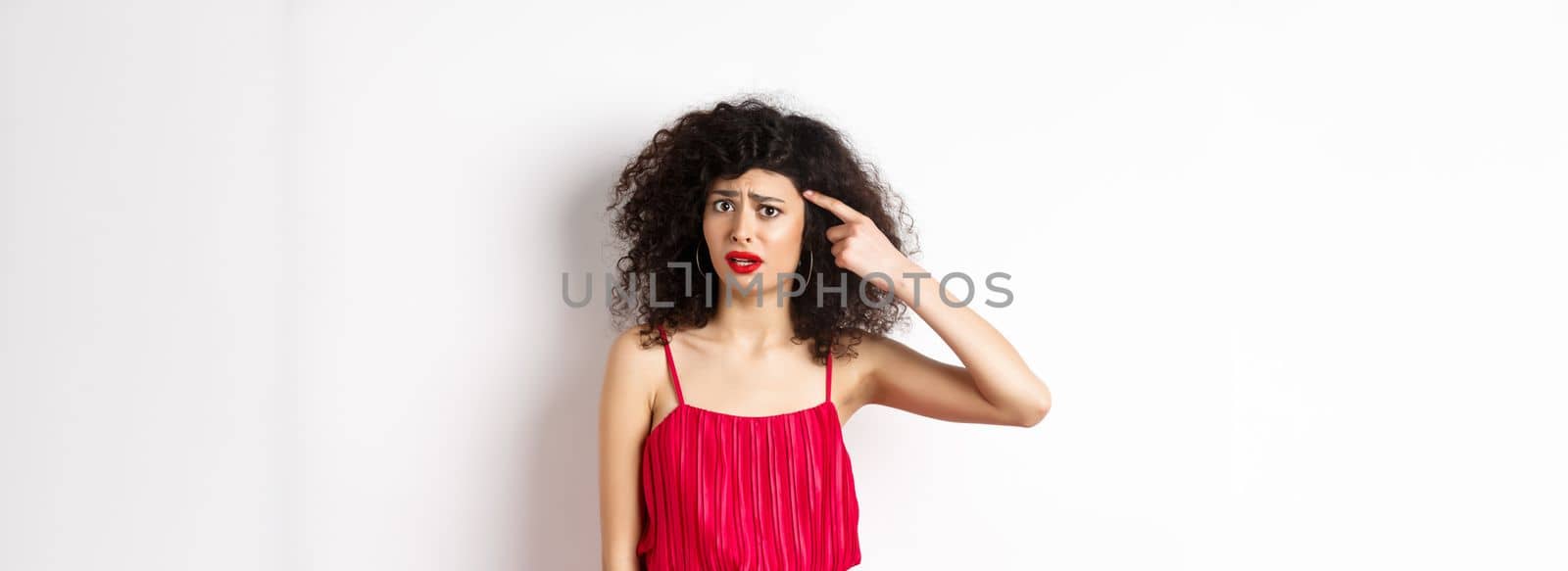 Shocked and confused caucasian woman in red dress scolding someone crazy, pointing finger at head and frowning, standing over white background by Benzoix