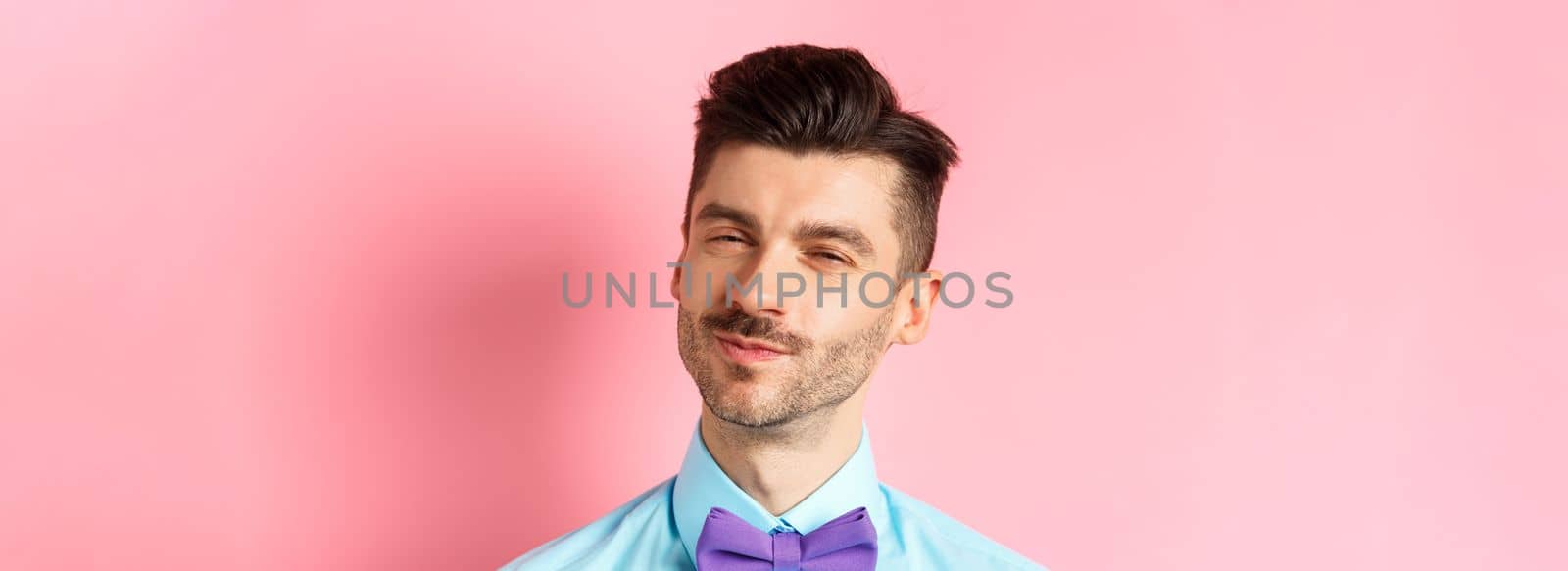 Close-up of smiling young guy with moustache, squinting intrigued, listening to something interesting, standing in bow-tie on pink background.