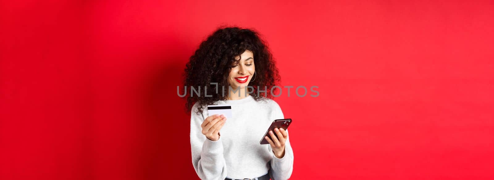 E-commerce and online shopping concept. Attractive caucasian woman paying for purchase in internet, holding smartphone and credit card, red background.