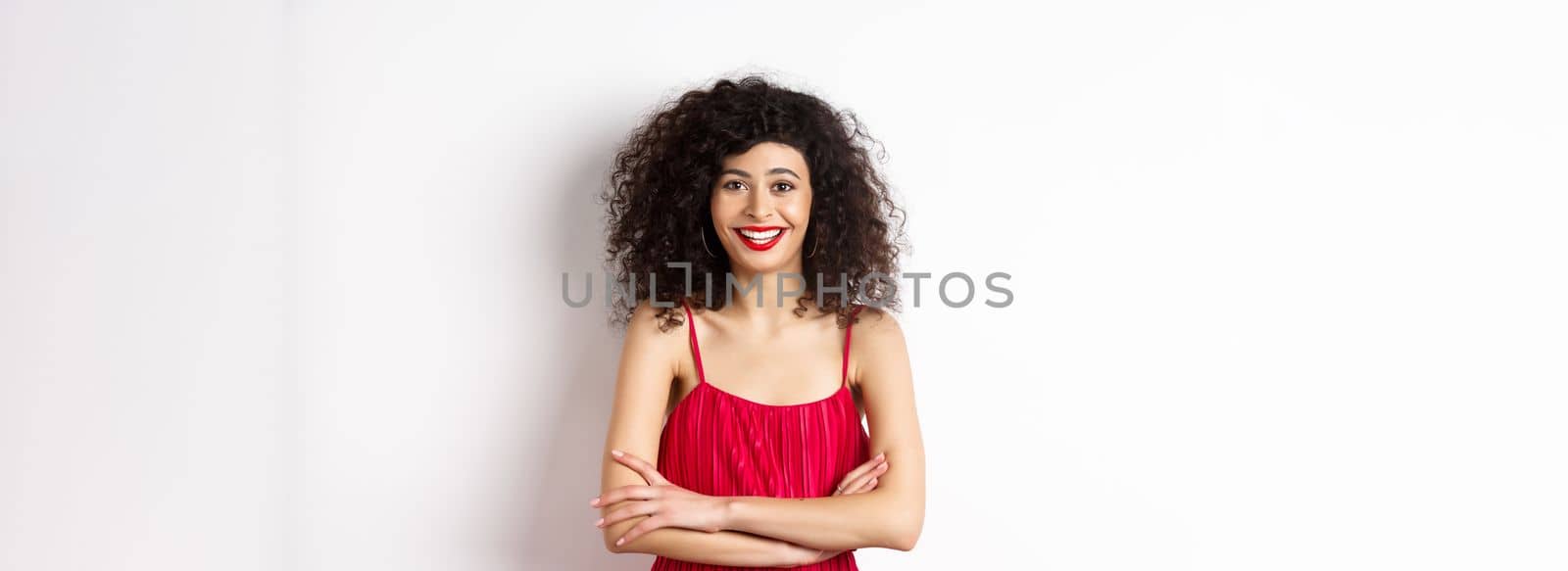 Elegant young woman in red dress with makeup, dressed-up for festive event, smiling happy at camera, standing over white background.