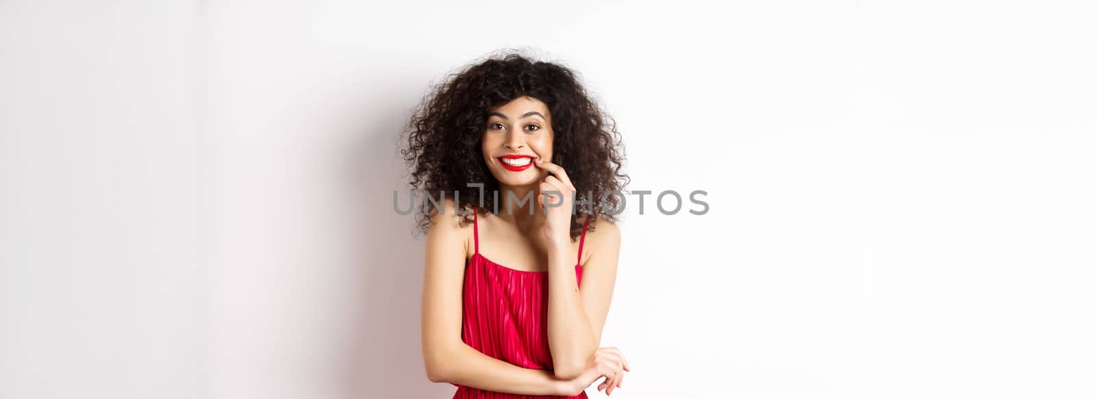 Excited beautiful woman in red dress, biting finger and smiling cute at camera, want something, standing on white background.