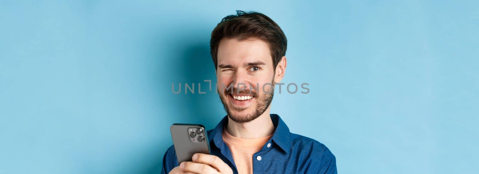 Close up of cheerful man winking and smiling, using mobile phone on blue background by Benzoix