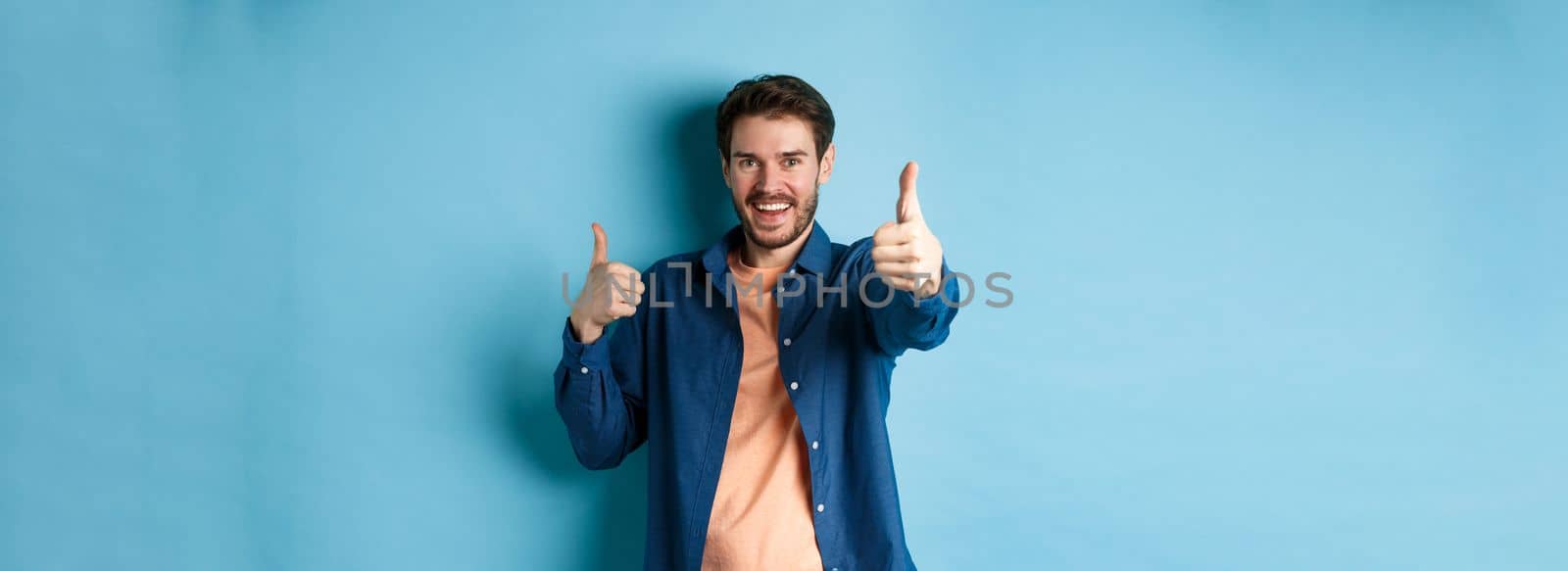 Positive smiling guy showing thumbs up and smiling, complimenting you, praising good job, well done gesture, standing on blue background by Benzoix