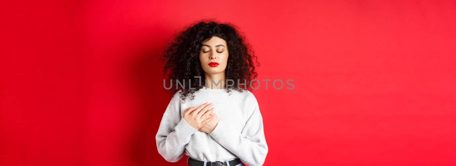 Image of calm young woman with curly hairsty, close eyes and holding hands on heart, keeping warm memories, feeling nostalgic, standing on red background by Benzoix