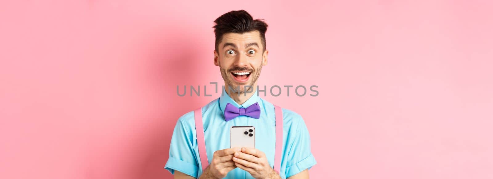 Online shopping. Happy man looking amazed after reading smartphone screen, smiling excited at camera, standing over pink background by Benzoix