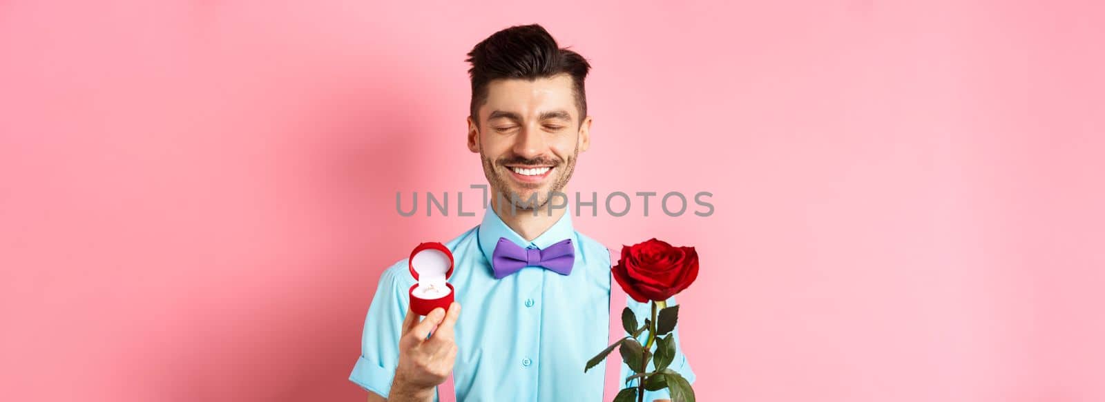 Valentines day. Cute boyfriend making wedding proposal, showing engagement ring in small box and red rose, express love, standing over pink background by Benzoix