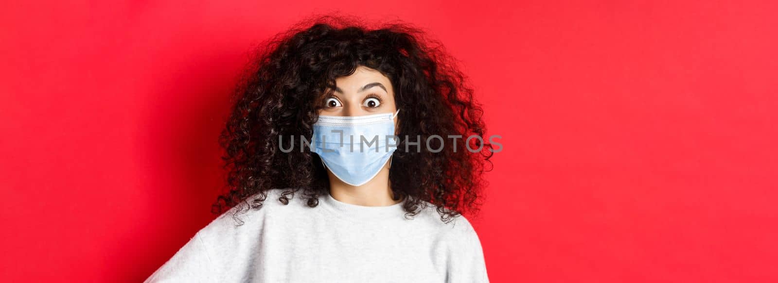 Covid-19 and pandemic concept. Close-up of surprised woman in medical mask pop eyes at something awesome, checking out promo, standing on red background.