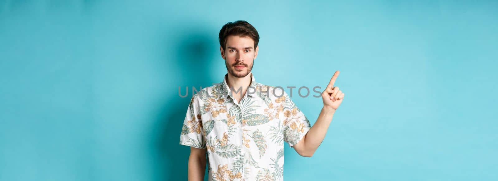Portrait of handsome bearded guy on vacation, wearing summer shirt and pointing finger left at logo, standing on blue background.