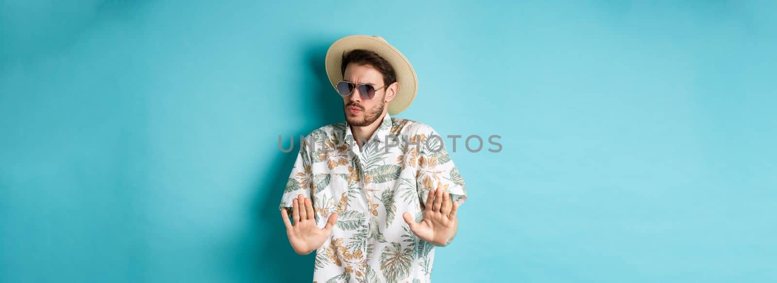 Alarmed tourist asking to stay away, step back from something cringe, showing rejection gesture, standing in straw hat and hawaiian shirt, blue background.