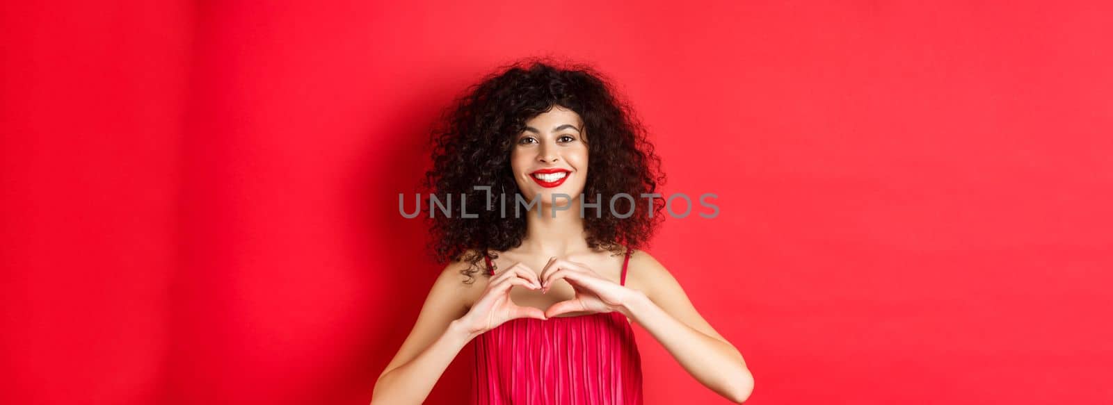 Valentines day. Romantic girl with curly hairstyle in evening dress, smiling and showing heart sign, say I love you on lovers holiday, standing over red background by Benzoix