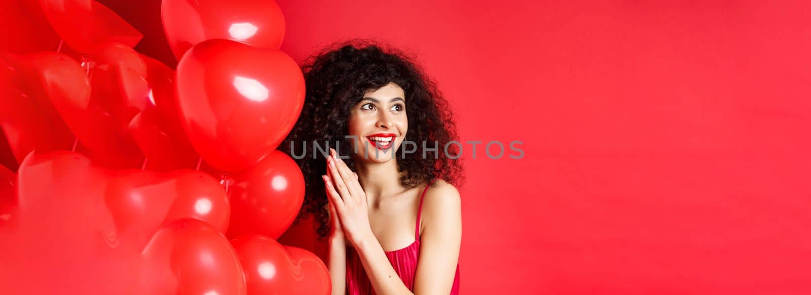Excited beautiful woman with curly hair, standing near heart balloons and rubbing palms together, expect good deal or relish, red background.