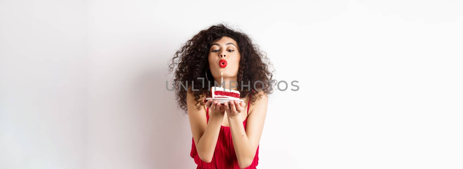 Holidays and celebration concept. Romantic woman in red dress celebrating birthday, blowing out candle on b-day cake and making wish, standing over white background.